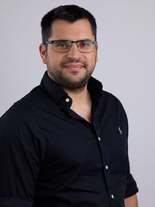 ד"ר לויטס גבר מחייך עם משקפיים בחולצה שחורה מעומדת. הרקע הוא צבע אפור בהיר, מה שמדגיש את הדמות. התמונה מתאימה לתחום של אסתטיקה רפואית.